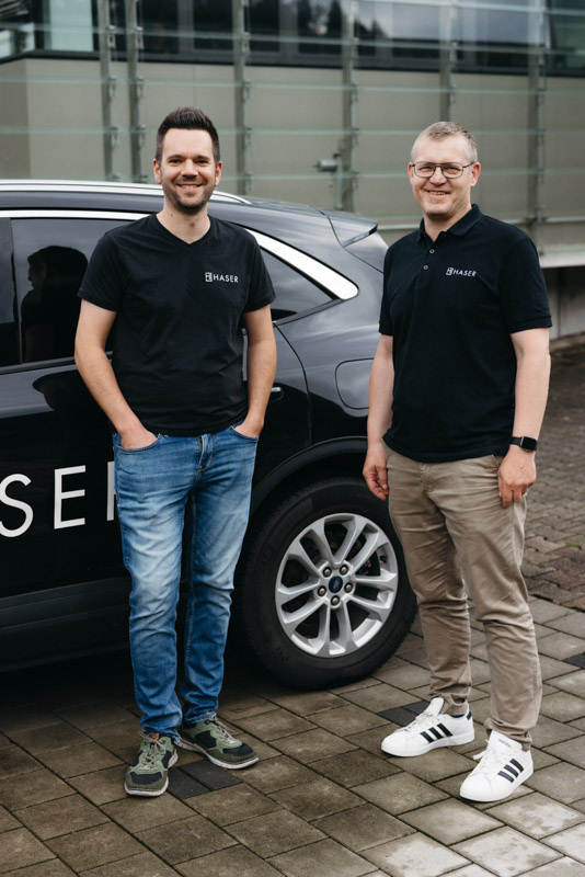 Zwei lächelnde Männer in Haser-T-Shirts stehen neben einem Auto. Erfolgreiche Karriere.