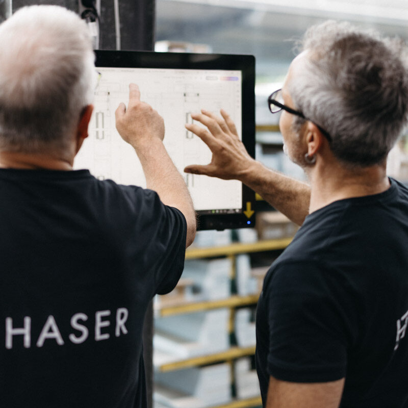 Zwei Kollegen arbeiten gemeinsam an einem Computerbildschirm an einem Projekt. Auf der Rückseite eines Shirts steht „HASER“.