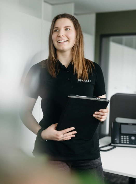 Lächelnde Frau in einem schwarzen HASER-Poloshirt, hält ein Klemmbrett. Karrierechance.
