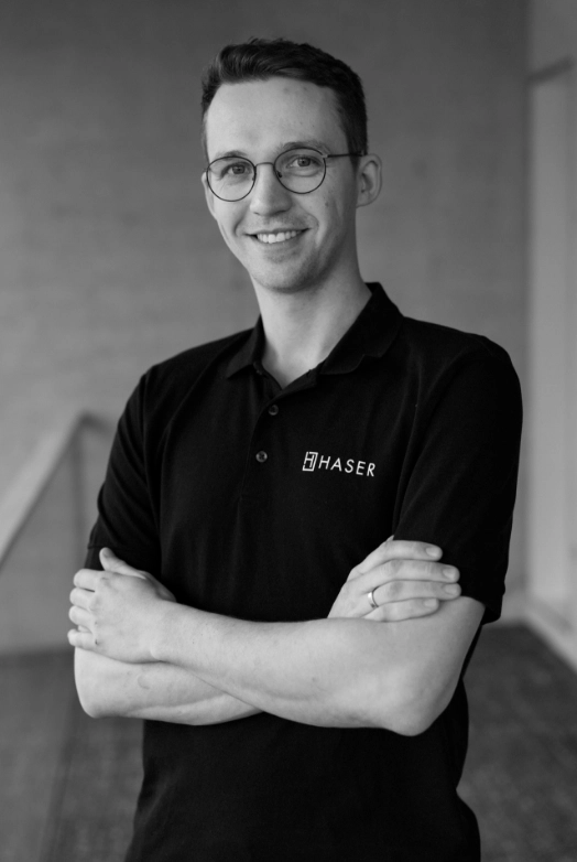 http://Porträt%20eines%20Mannes%20mit%20verschränkten%20Armen,%20der%20ein%20schwarzes%20Haser-Poloshirt%20trägt.
